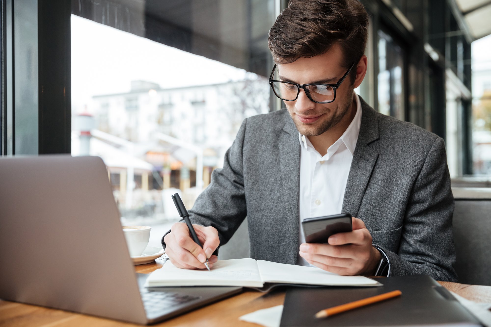 smiling-businessman-eyeglasses-sitting-by-table-cafe-with-laptop-computer-while-using-smartphone-writing-something-2